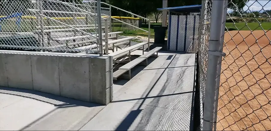 Concrete walkway and retaining wall in front of aluminum bleachers beside a ballfield fence