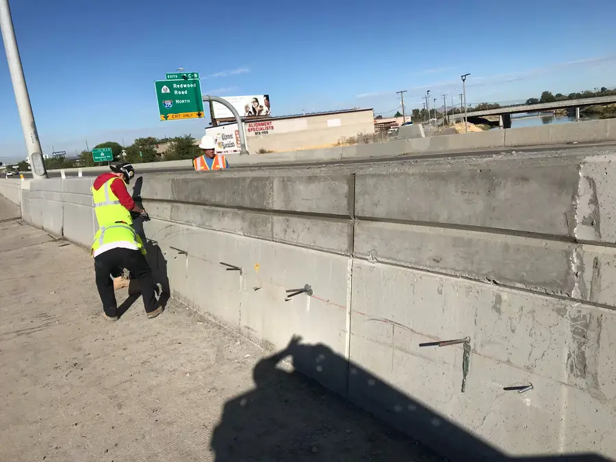 Worker drilling into a concrete barrier on a highway bridge, with steel dowels set along the wall