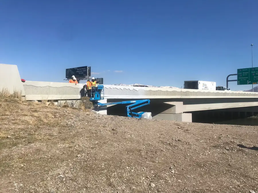 Workers in a boom lift at the bridge parapet over a channel, with highway signs and mountains beyond