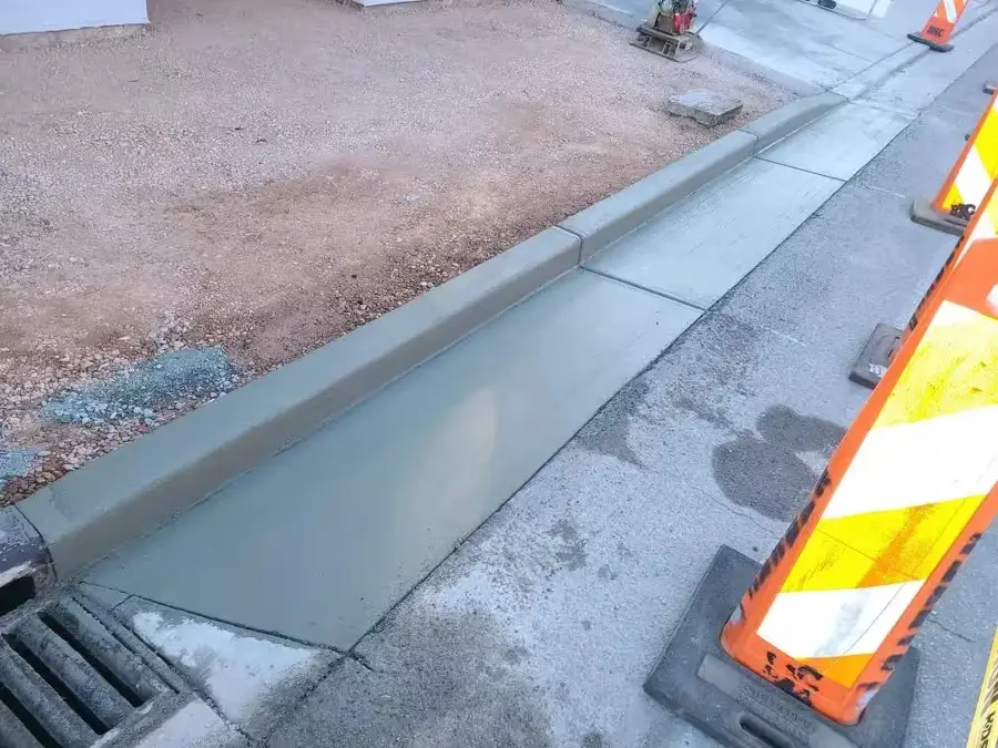 Freshly finished concrete curb and gutter meeting a storm drain grate, behind orange traffic barricades