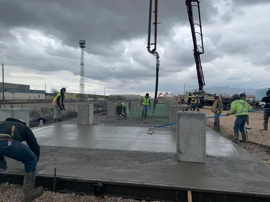 B Hansen Construction crew placing and screeding a concrete slab around foundation piers, with a concrete pump boom overhead