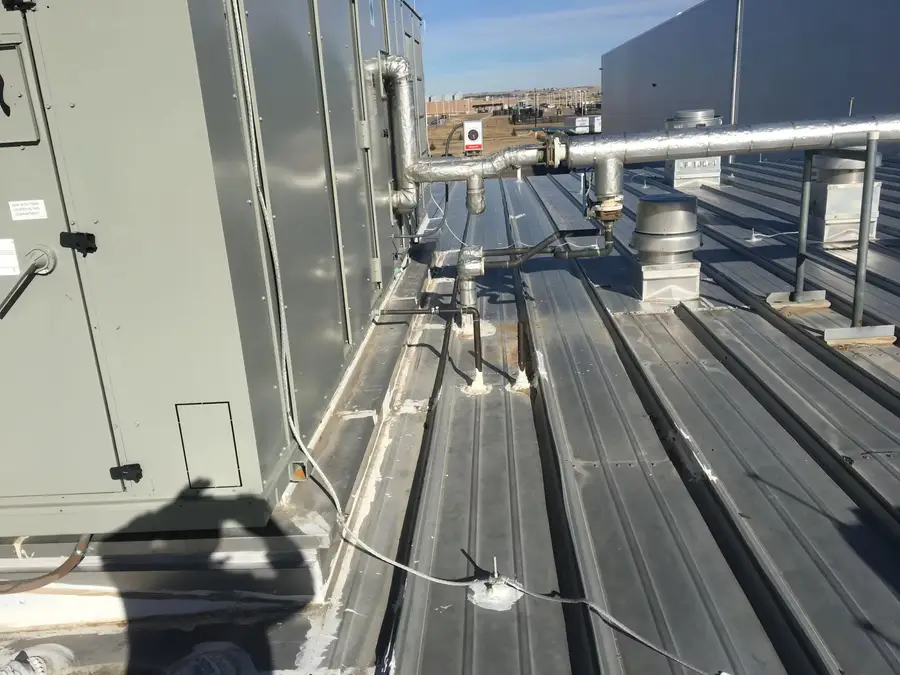 Insulated pipes and vents crossing a standing seam metal roof beside a rooftop HVAC unit