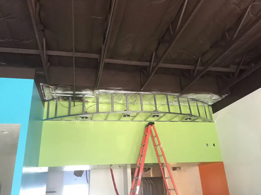 Curved metal framing for a lighted soffit above a green wall, with a ladder in place