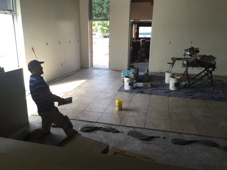 Tile setter kneeling with a trowel while laying floor tile, with a wet tile saw set up nearby