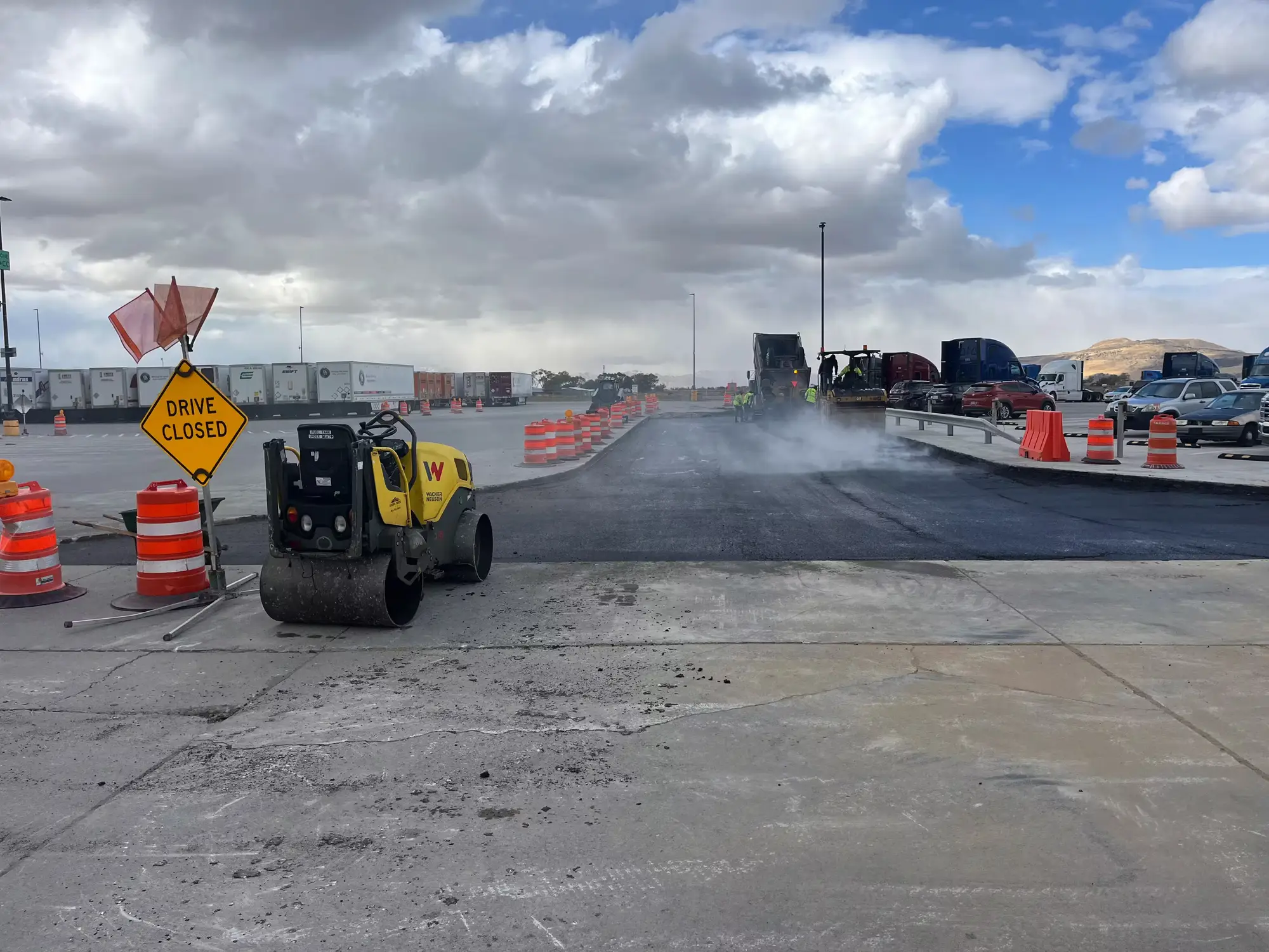 A roller compacting fresh asphalt behind traffic barrels and a Drive Closed sign