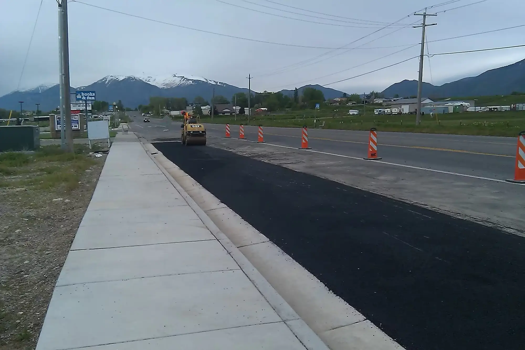 New concrete sidewalk and fresh asphalt along a highway, with a roller working behind traffic cones and snow-capped mountains beyond